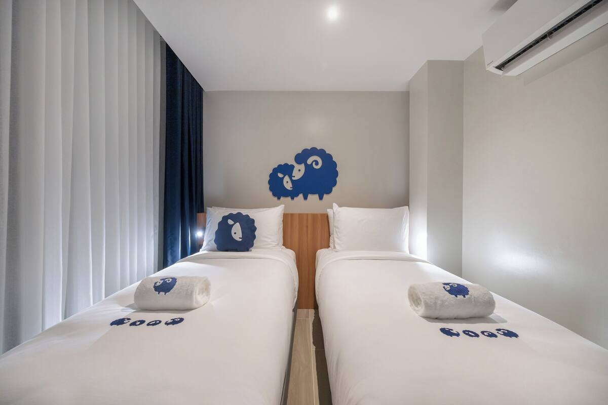 Une chambre d’hôtel avec deux lits, chacun agrémenté d’un coussin à motif de mouton bleu et d’une serviette ornée du même motif.