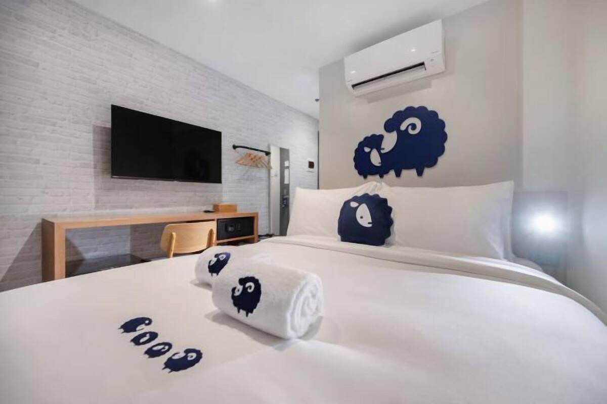Une chambre d’hôtel moderne avec un mur en briques blanches, un téléviseur à écran plat fixé au-dessus d’un bureau en bois, un lit avec une literie blanche et des coussins décoratifs bleu foncé, et un climatiseur mural.