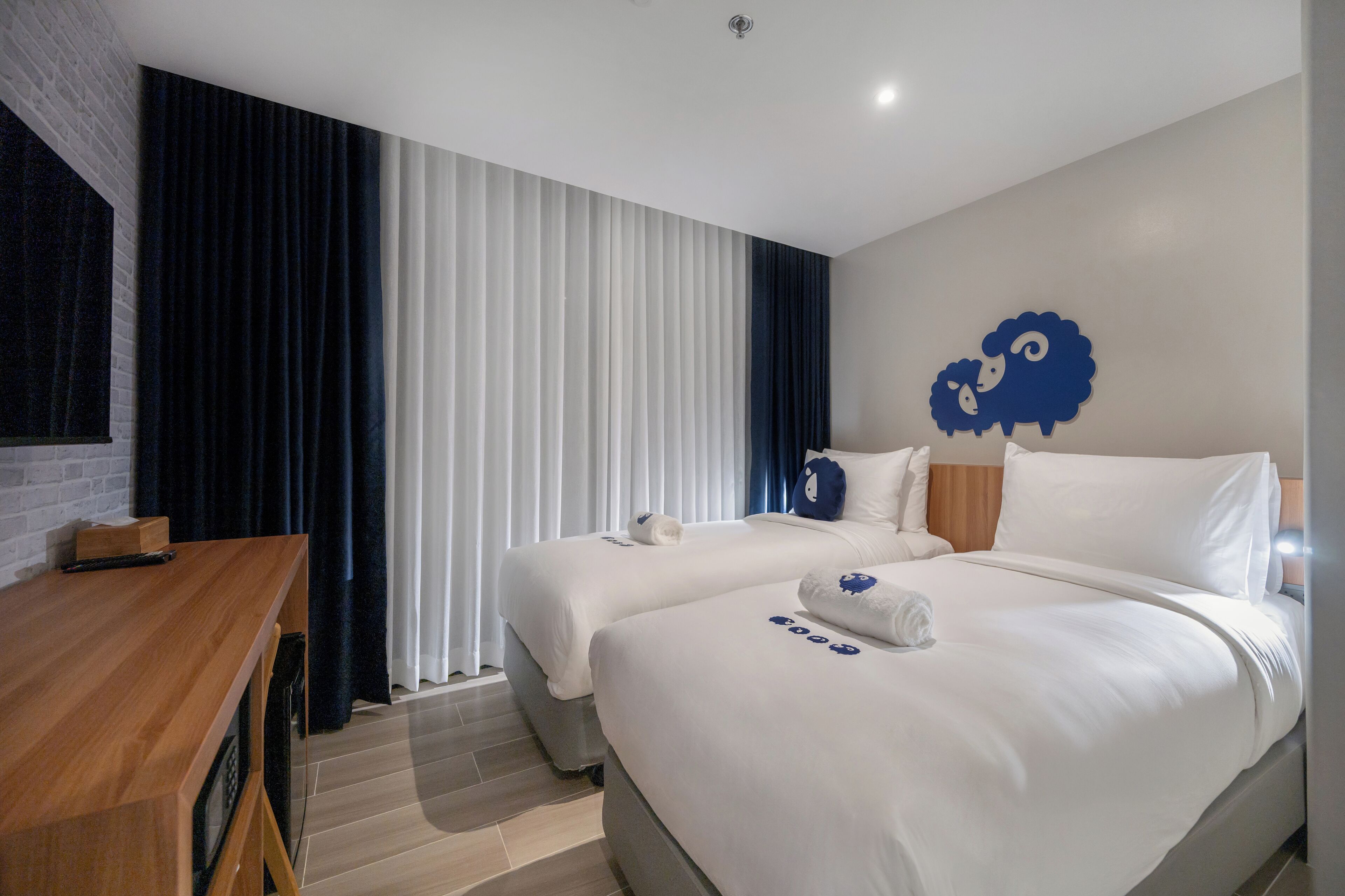 A hotel room with two beds, a wooden desk, a large window with blinds, and a wall-mounted sheep decoration.