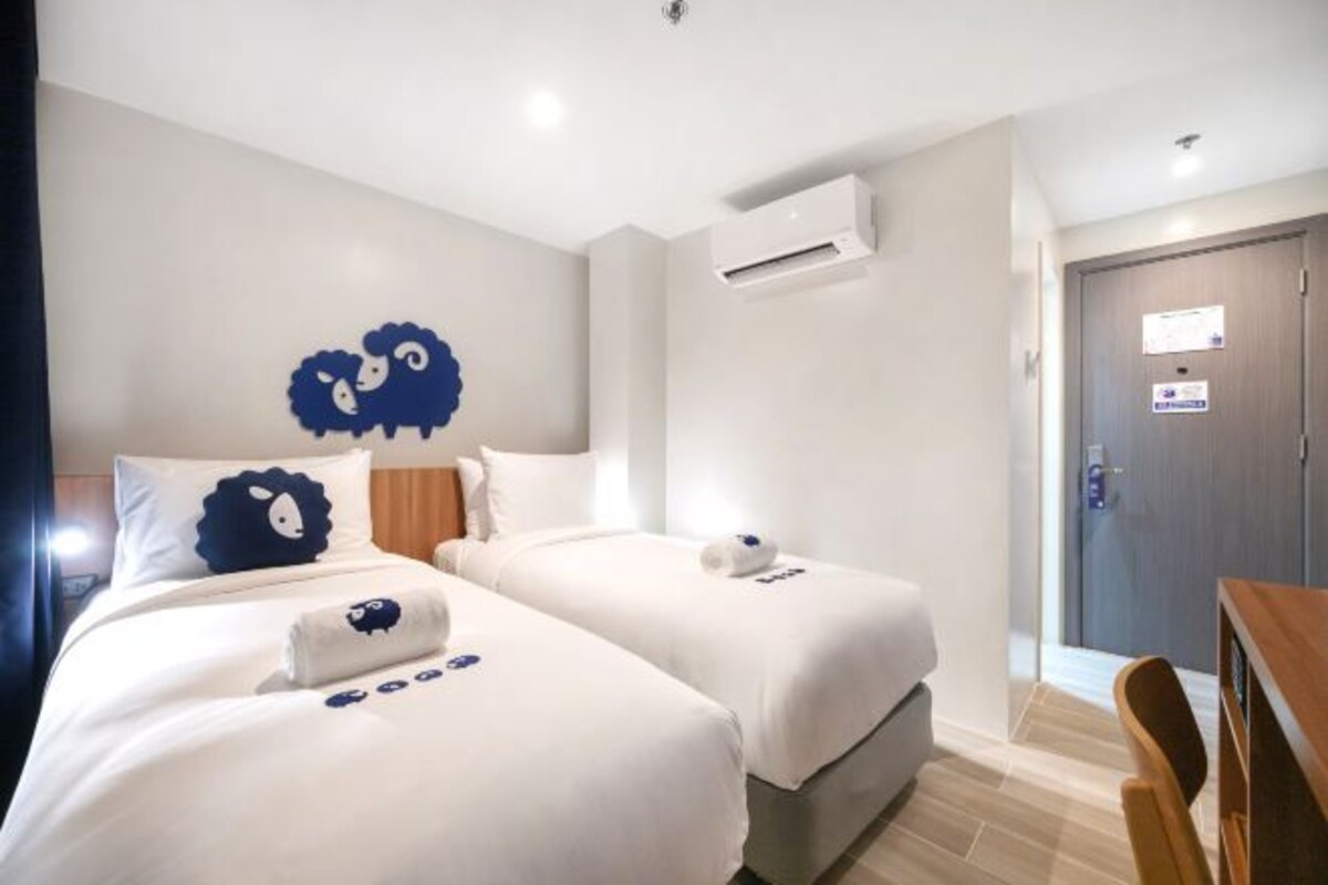 Une chambre d’hôtel avec deux lits, une tête de lit en bois, un appareil de climatisation, une porte avec une pancarte et une petite table.