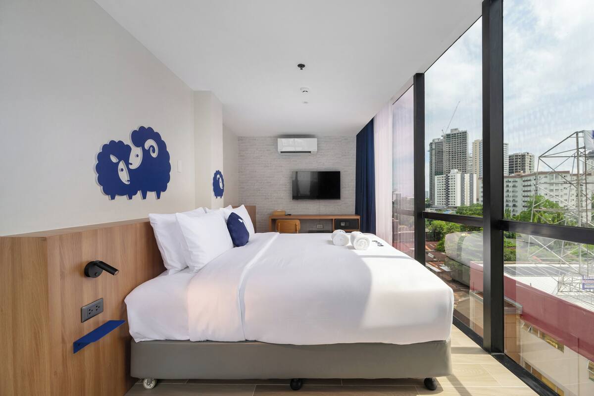 Une chambre d’hôtel moderne dotée d’un grand lit, d’un bureau et offrant une vue sur la ville grâce à de grandes fenêtres du sol au plafond.