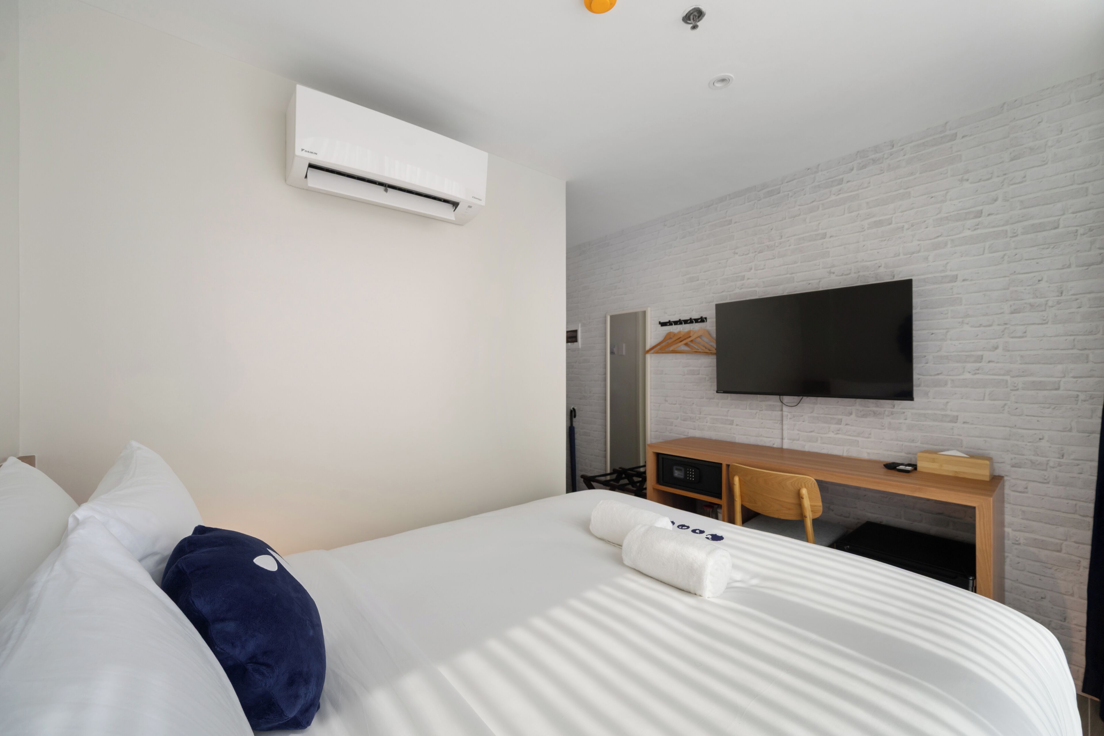A hotel room with a bed, white walls, a flat-screen TV mounted on a brick wall, a wooden desk, and an air conditioning unit.
