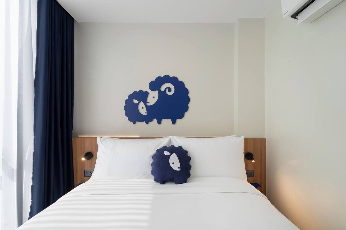 Une chambre d’hôtel avec un lit, deux oreillers, une œuvre d’art représentant des moutons accrochée au mur, une tête de lit en bois et une table de chevet avec une lampe.