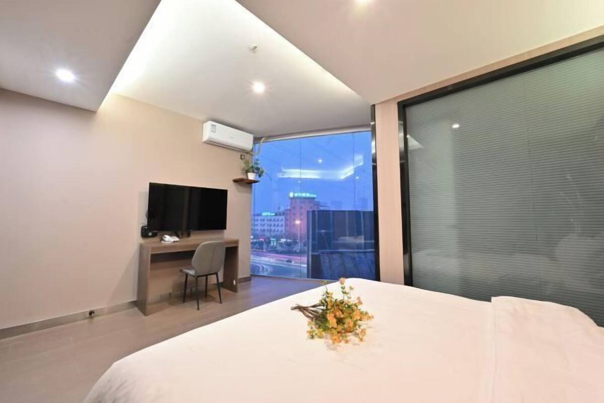 Habitación de hotel con cama, escritorio, silla, televisor y un amplio ventanal con vista a la ciudad.