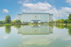 A large, modern building with a symmetrical facade, a central entrance, and a reflection in the water in front.