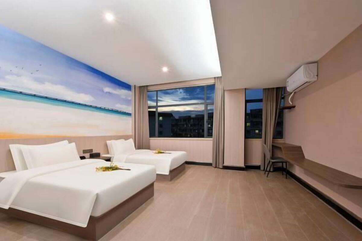 Habitación de hotel con dos camas, un ventanal con vistas a la ciudad, un cuadro de una playa en el techo y un televisor montado en la pared.