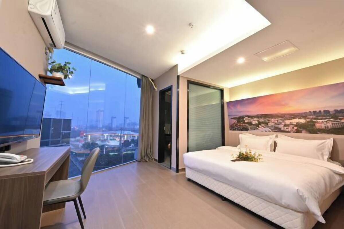 Una habitación de hotel moderna con una cama grande, un escritorio con silla, un ventanal con vista a la ciudad y un cuadro en la pared.