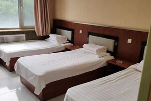 A hotel room with three single beds, wooden headboards, a window with curtains, and a radiator.