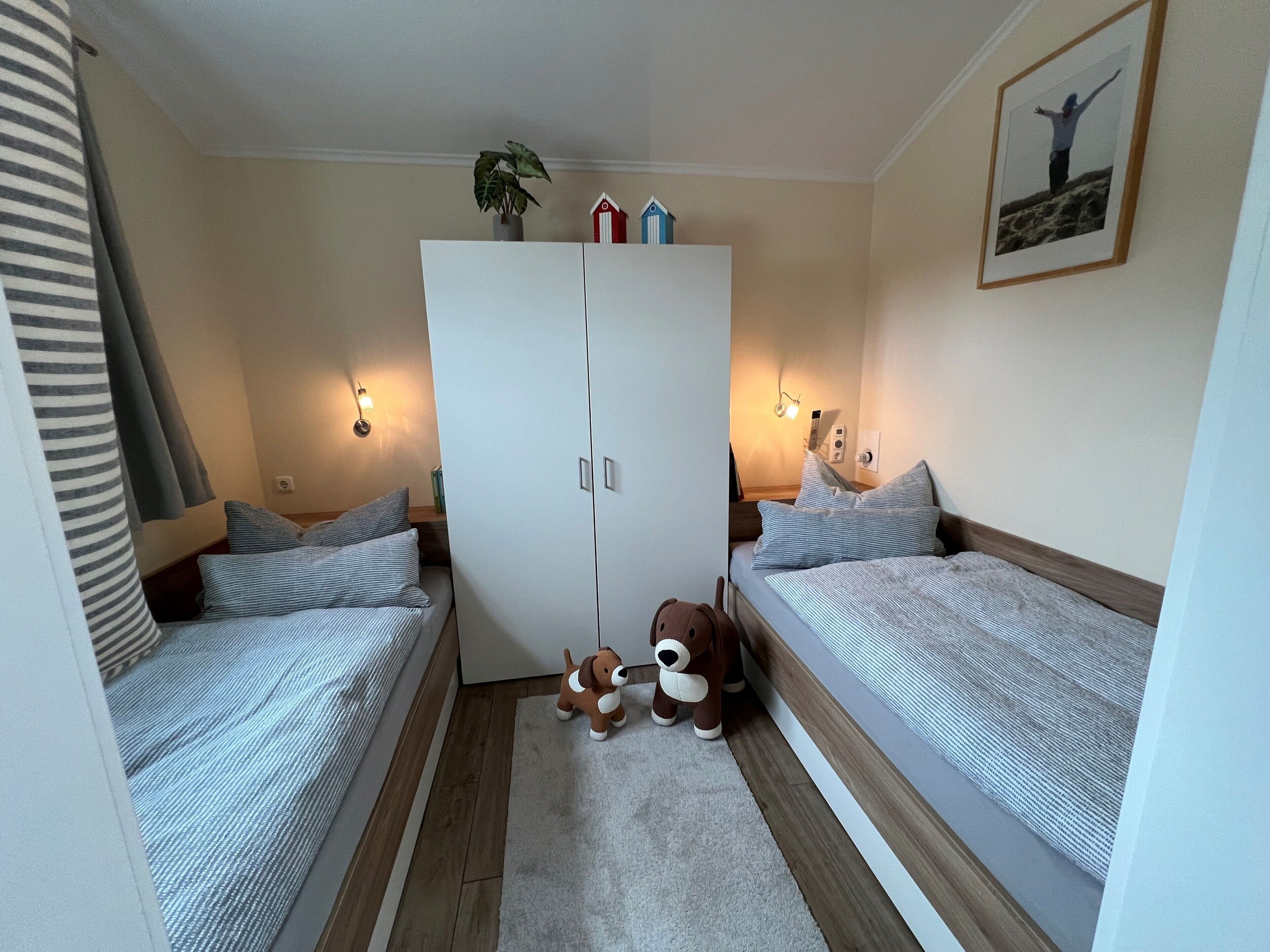 Um quarto com duas camas de solteiro, um guarda-roupa, uma mesinha, uma luminária, um urso de pelúcia e um quadro na parede.