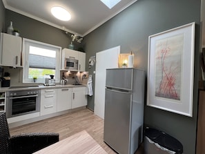Een moderne keuken met een koelkast, magnetron en oven, een eethoek met tafel en stoelen, en een raam met jaloezieën.