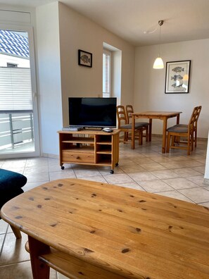 A room with a TV on a wooden stand, a dining table with chairs, a window with blinds, and a ceiling light.