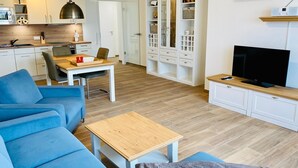 A modern living room with a blue sofa, a wooden coffee table, a TV, and a kitchen area with cabinets and a microwave. - Apartment 'Duhner Allee 10, Litfaß 17' with Balcony and Wi-Fi (Cuxhaven)