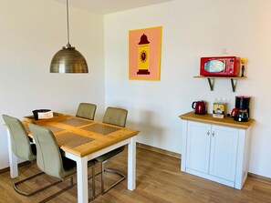 Een eettafel met stoelen, een hangende hanglamp, een magnetron, een koffiezetapparaat en een kast met een plank.