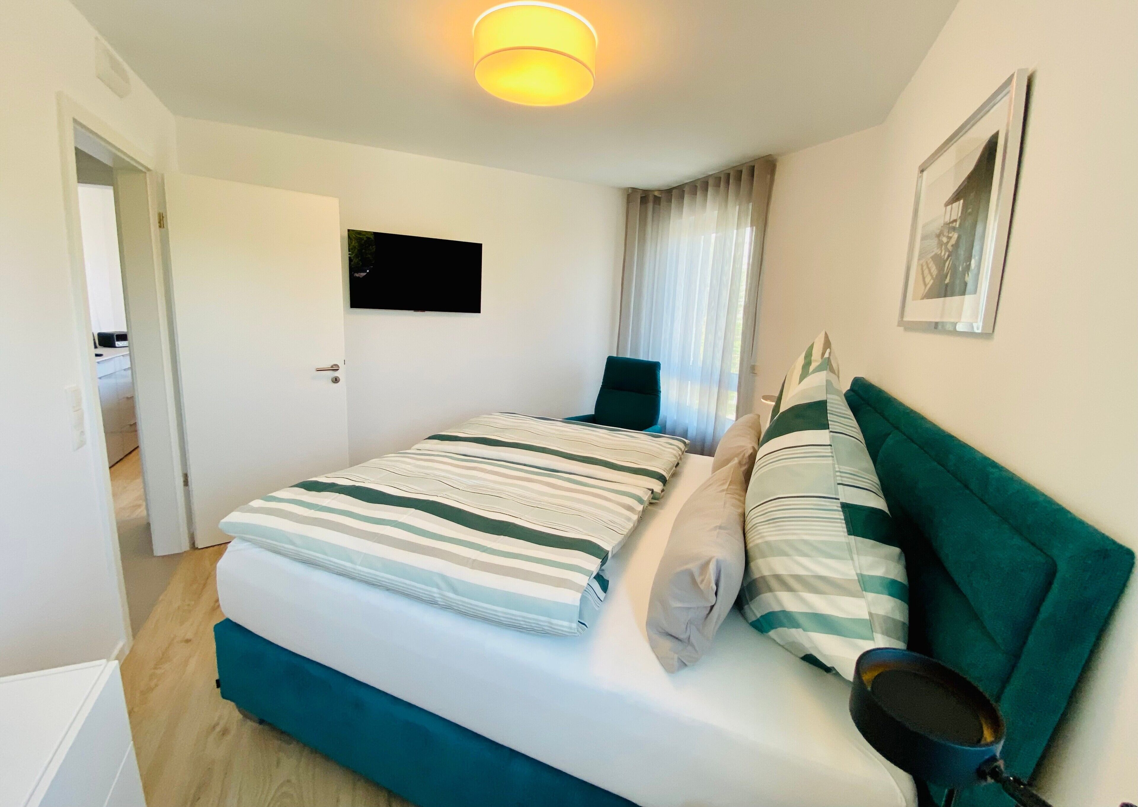 Ein ordentlich bezogenes Bett mit gestreifter Bettwäsche, ein türkisfarbenes Kopfteil, ein Flachbildfernseher über dem Bett, eine Tür zu einem anderen Raum und ein gerahmtes Bild an der Wand.