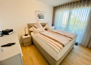Une chambre d’hôtel avec un lit, une table de chevet dotée d’une radio et d’une télécommande, une lampe et une fenêtre avec des rideaux.