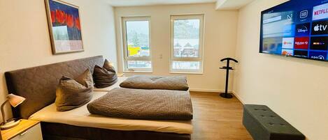 A bedroom with a bed, bedside table, lamp, artwork, windows, and a TV displaying streaming services.