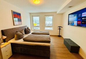 Ett sovrum med en säng, ett nattduksbord, en lampa, konstverk, fönster och en tv som visar strömnings­tjänster.