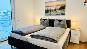 En dubbelsäng med en sänggavel, två sängbord med lampor, ett stort fönster och en tavla som föreställer en strandvy.