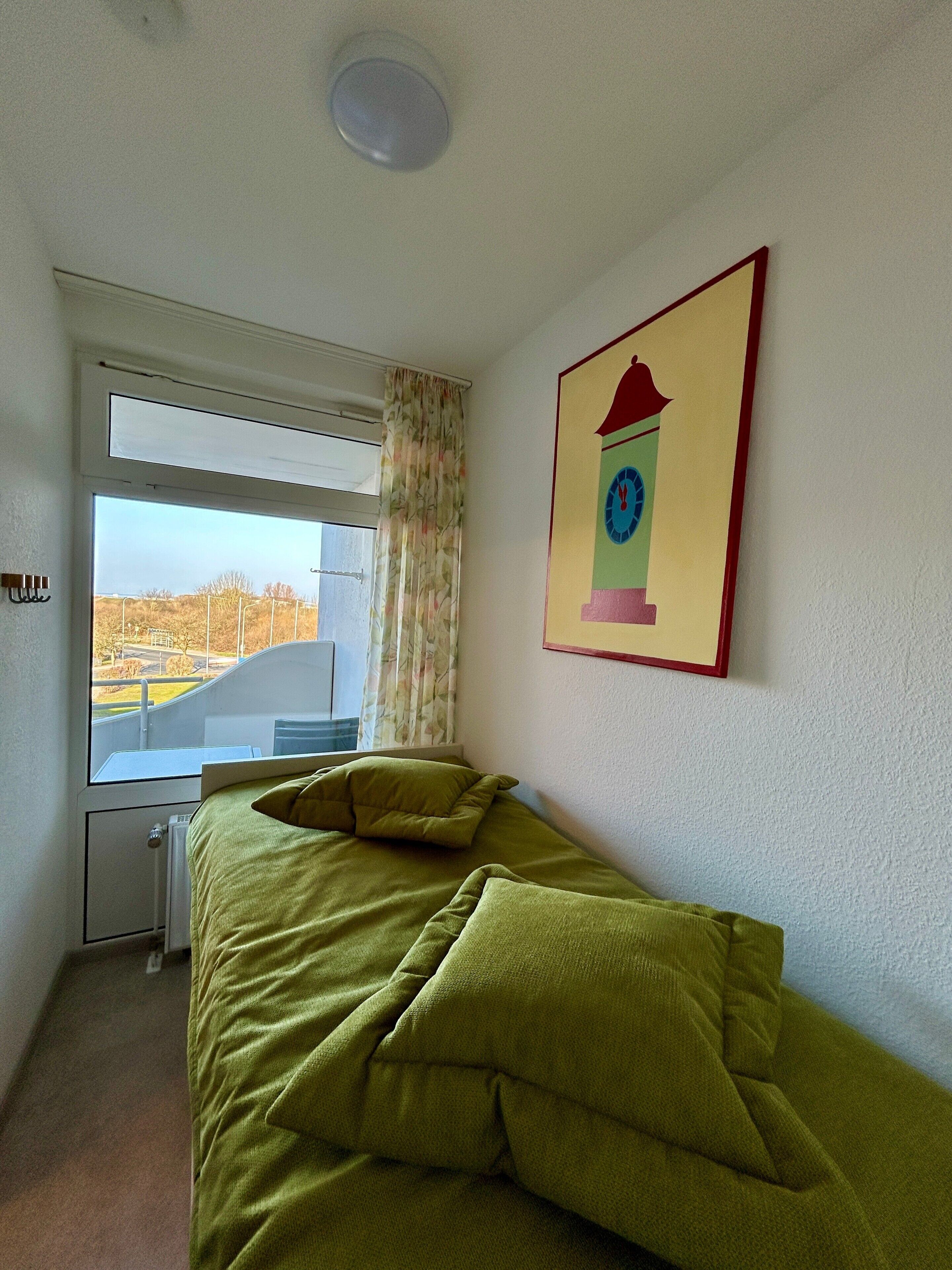 Ein Zimmer mit Bett, Fenster mit Blick, Heizkörper und einer Wanduhr.