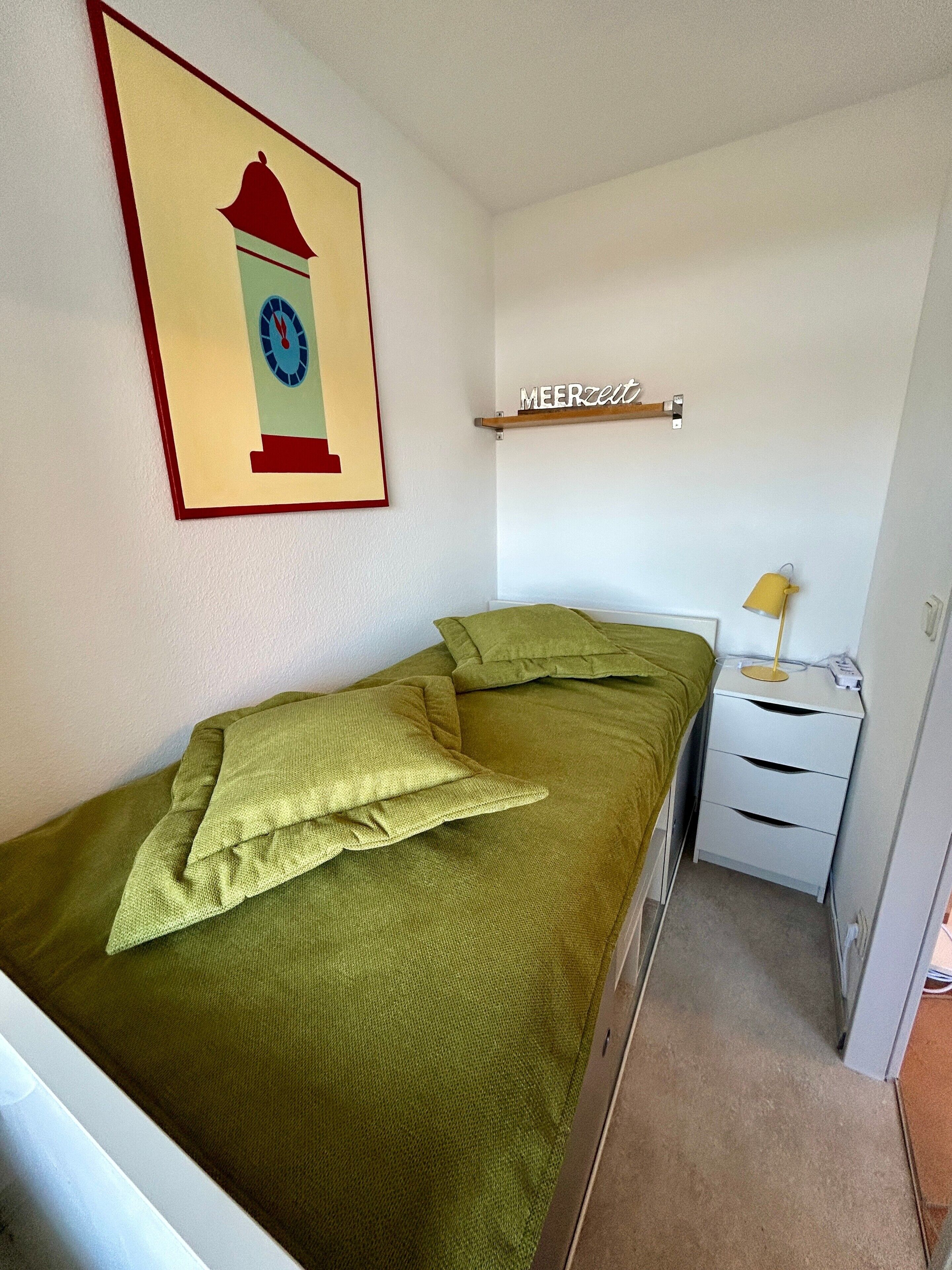 Ein kleines Zimmer mit einem Einzelbett, einer grünen Bettdecke, einem Wandposter mit einer Uhr, einem Nachttisch mit Lampe und einem Regal mit einem Schild.