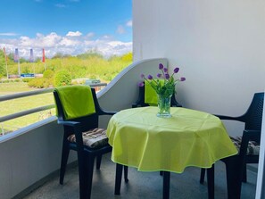 Un balcone con tavolo e sedie, un vaso con fiori viola e vista su vegetazione e bandiere.