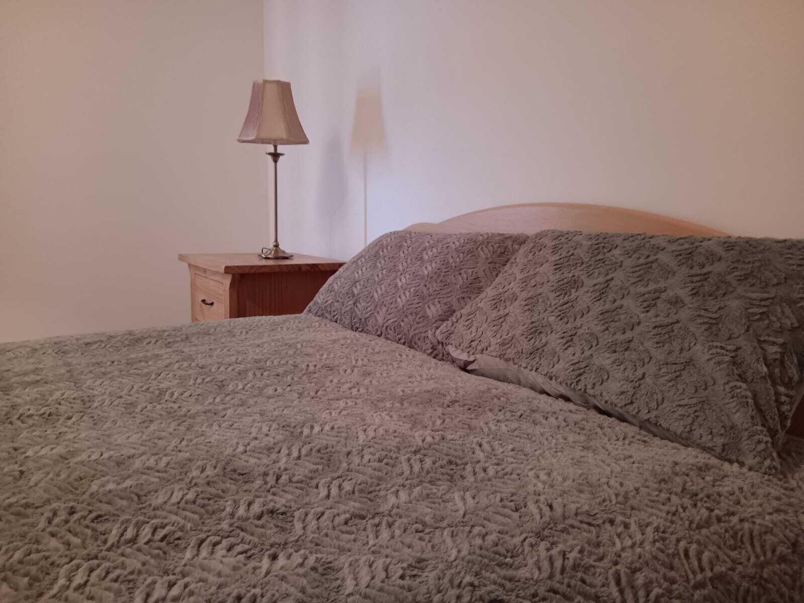 Una cama bien hecha con una colcha estampada y almohadas, una mesita de noche de madera con una lámpara, y una pared lisa al fondo.