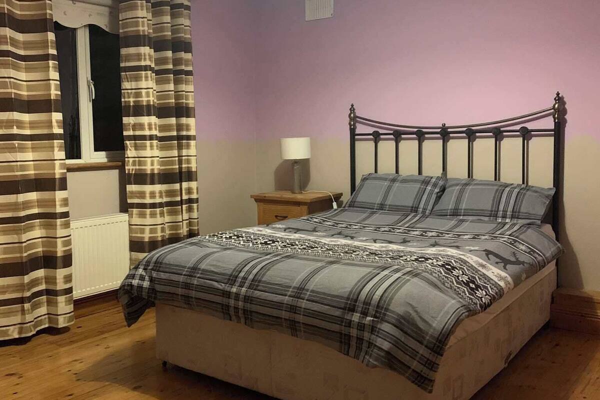 A bedroom with a bed, wooden floor, curtains, a nightstand, and a lamp.