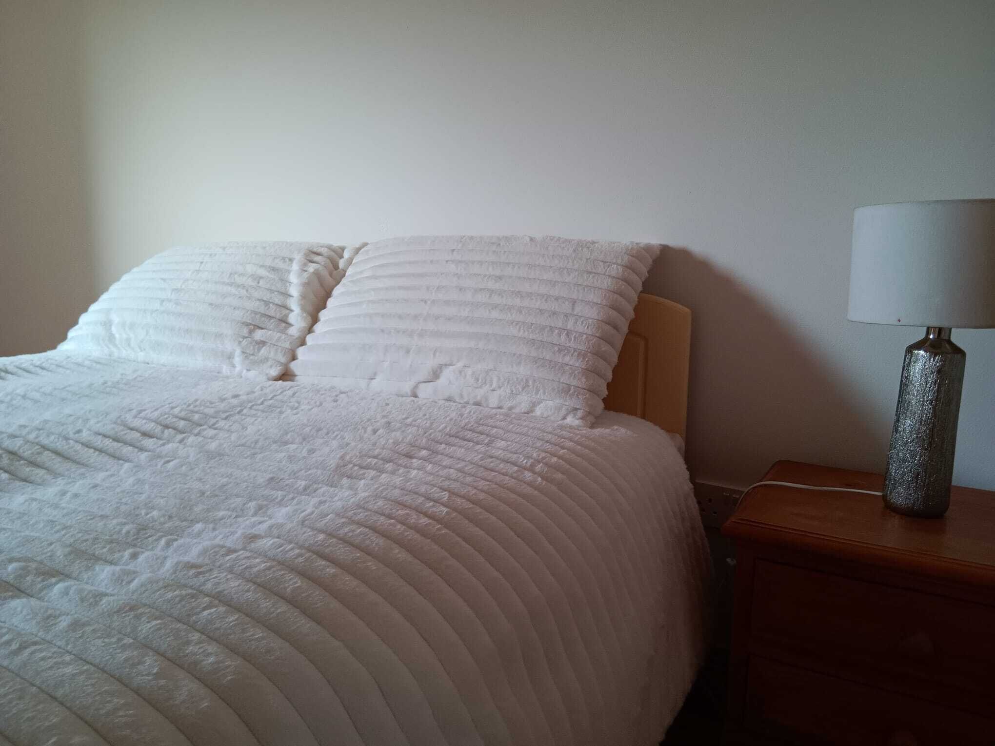 Una cama bien hecha con edredón blanco y almohadas, una mesita de noche de madera con lámpara y una pared lisa al fondo.