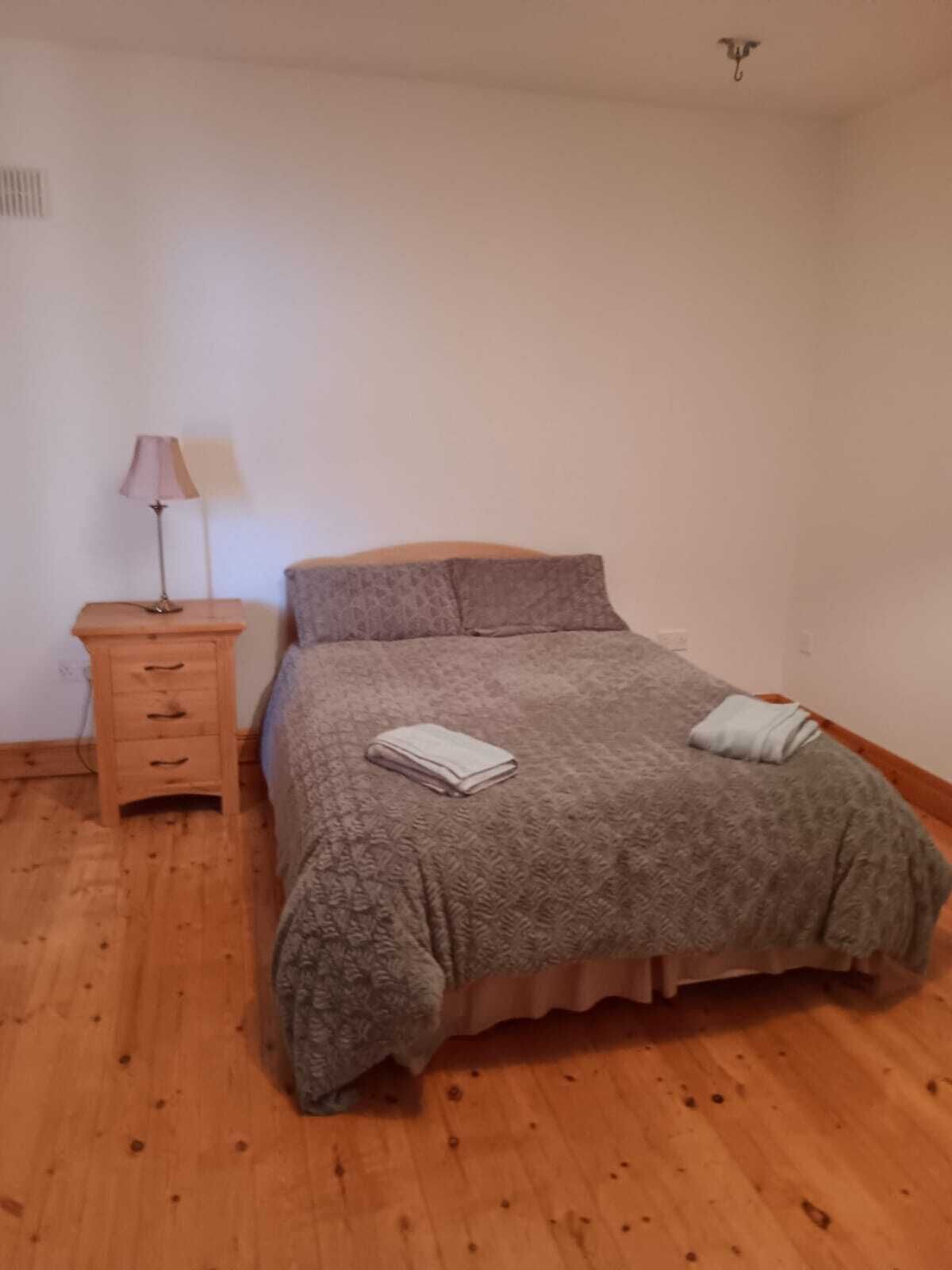 A bedroom with a bed, a nightstand, a lamp, and a wooden floor.
