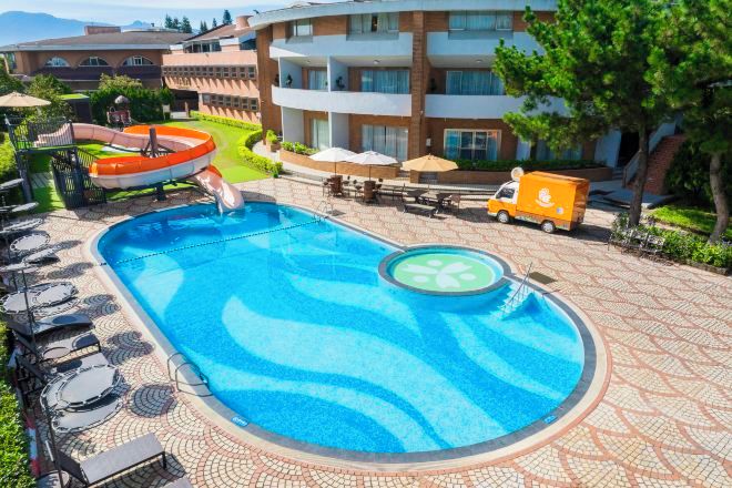A swimming pool with lounge chairs and a lifeguard stand, surrounded by a tiled walkway and a building with balconies.