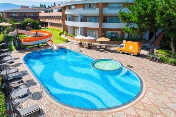 Una piscina con sillones de descanso y un puesto de socorrista, rodeada por un paseo cerámico y un edificio con balcones.