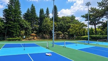 An outdoor tennis court with multiple courts, surrounded by trees and a clear sky.