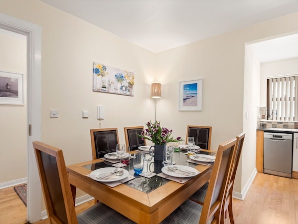 Un comedor con mesa de madera dispuesta para cuatro personas, rodeada de sillas, con obras de arte en las paredes y una cocina visible al fondo.