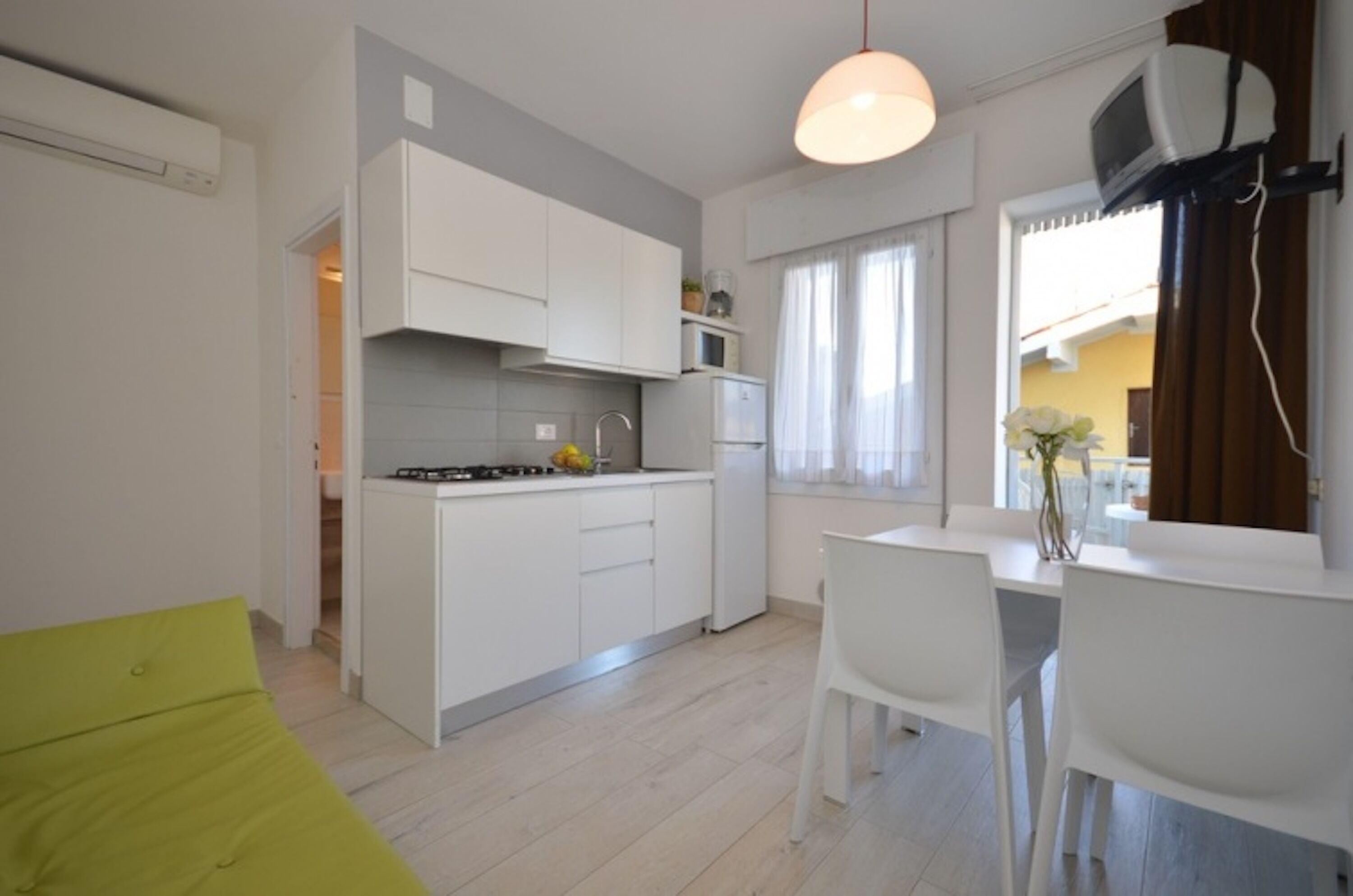 Een kleine keuken met witte kasten, een eethoek met een witte tafel en stoelen, en een raam met gordijnen.