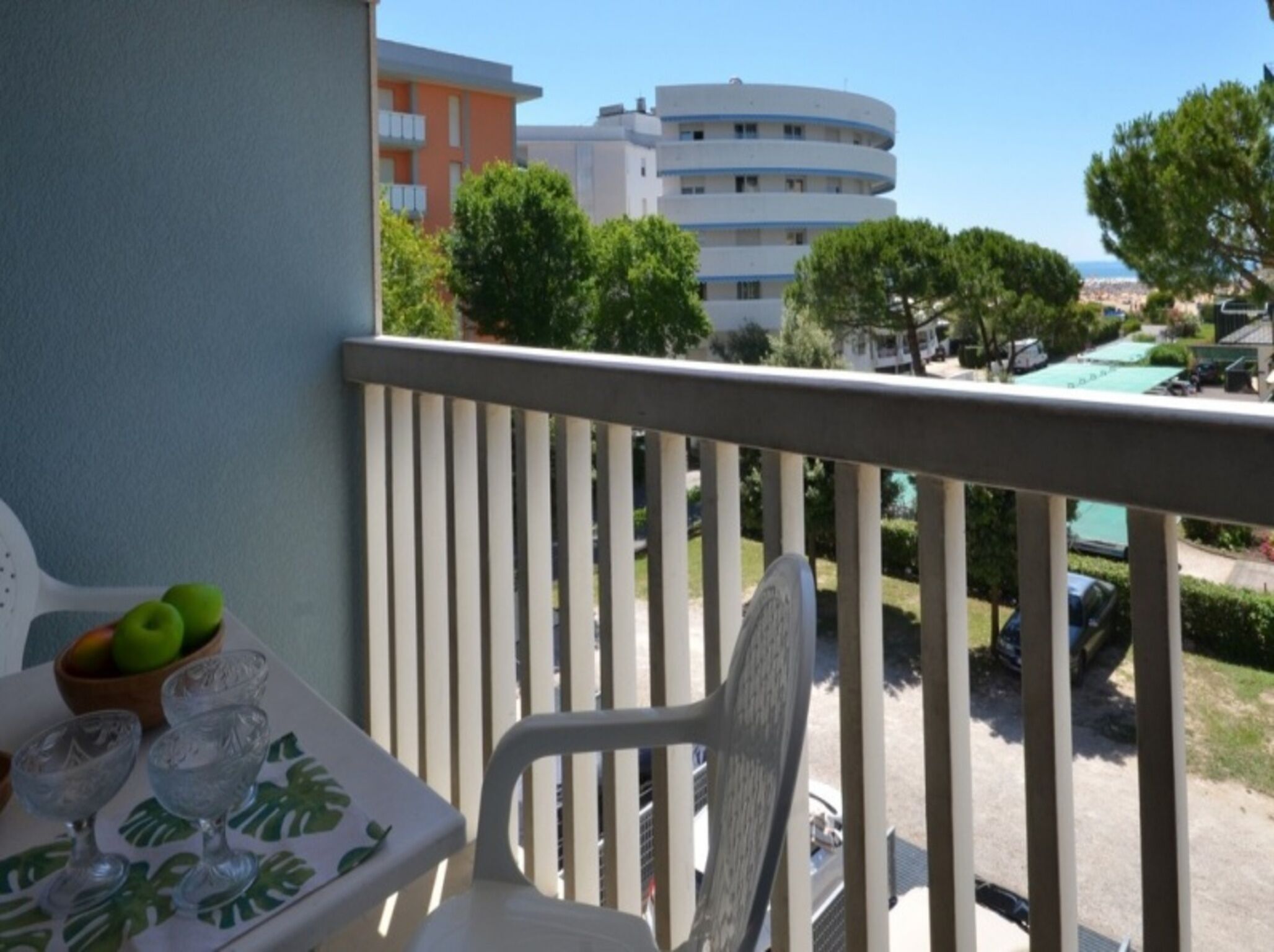 Un balcon avec une table, des chaises et un saladier rempli de fruits. En arrière-plan, on aperçoit des bâtiments et des arbres.