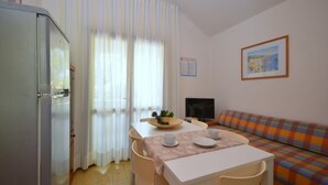 Una habitación pequeña y luminosa con una mesa de comedor, sillas, un sofá, un televisor y un refrigerador.