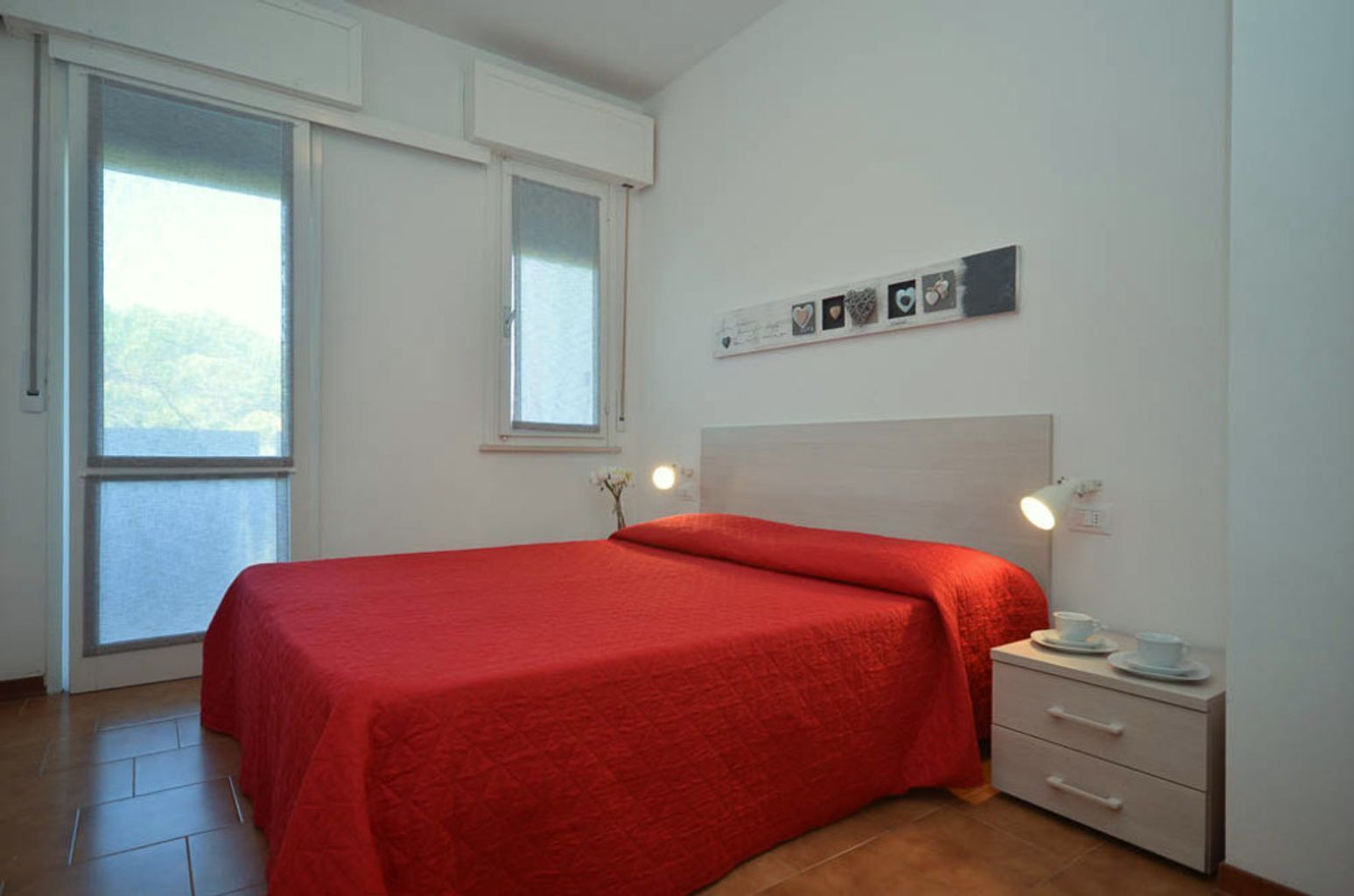 Een slaapkamer met een bed, nachtkastje, lamp, raam en een wand met foto’s.
