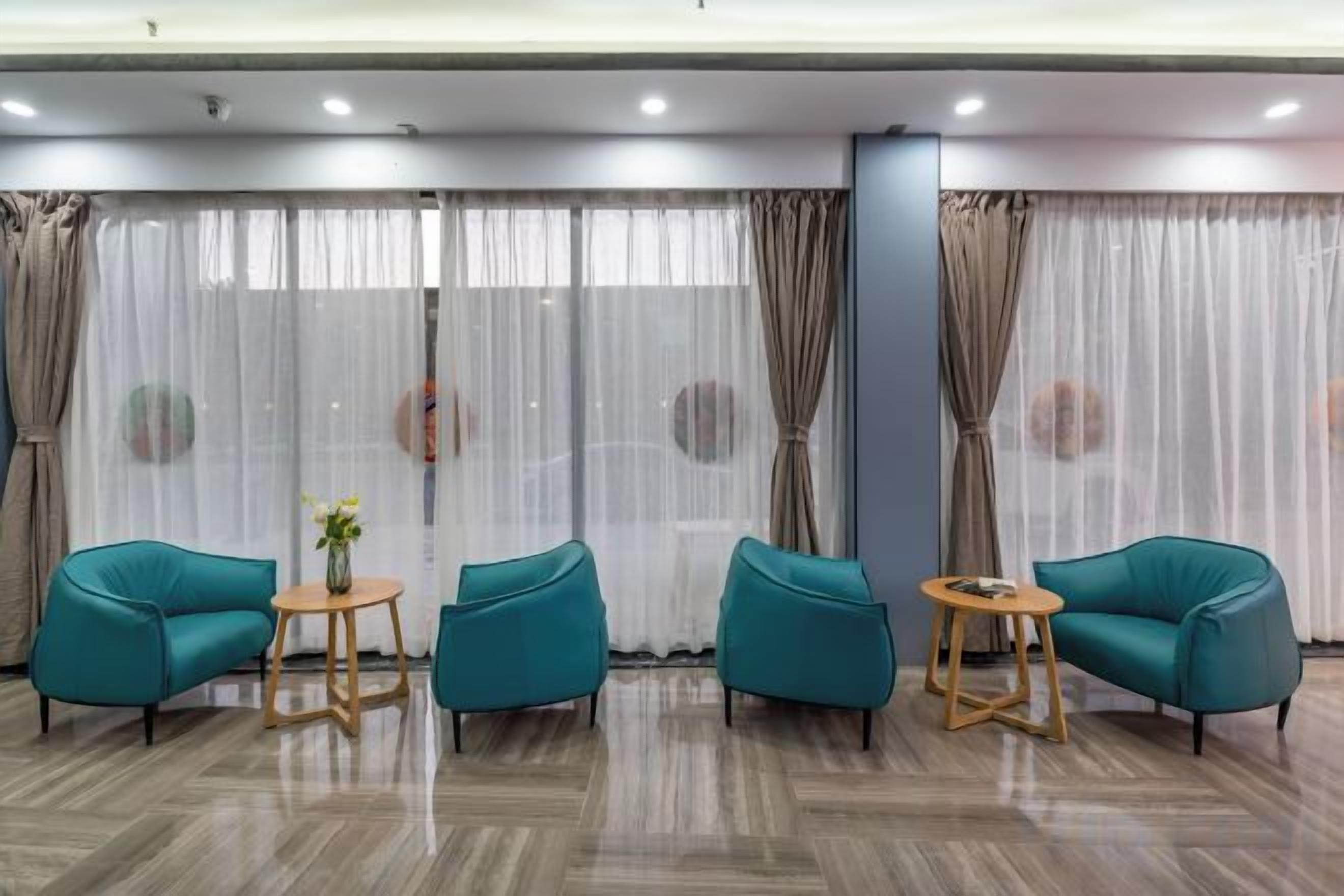 A room with a seating area featuring two teal chairs and a small round table, a large window with sheer curtains, and a polished wooden floor.