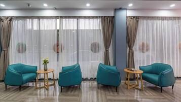 A room with a seating area featuring two teal chairs and a small round table, a large window with sheer curtains, and a polished wooden floor.