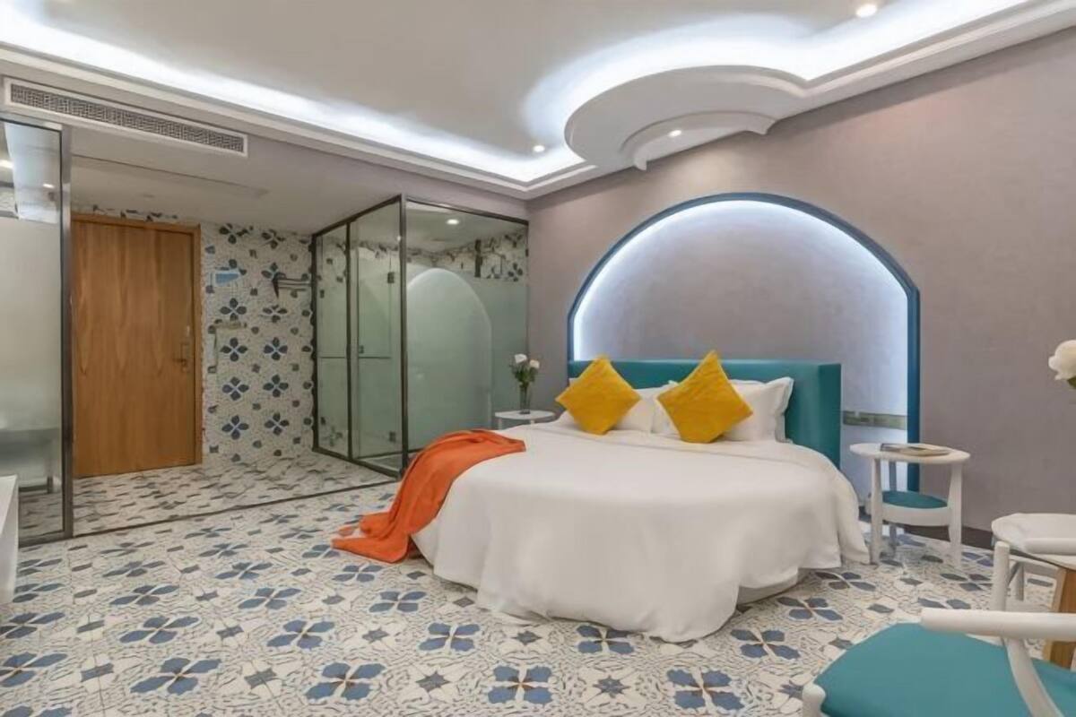 Ein modernes Hotelzimmer mit einem großen Bett, einer Duschkabine aus Glas und einer Deckenleuchte mit gebogenem Lampenschirm.