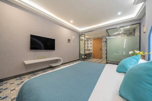 Una habitación de hotel con cama, televisión, baño con puerta de vidrio y una pequeña zona de estar.