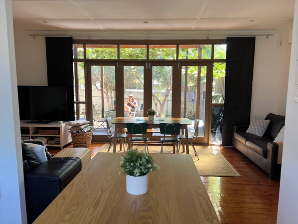 Una sala moderna con una mesa de comedor de madera, sillas verdes, un sofá de cuero negro y un televisor sobre un soporte blanco.