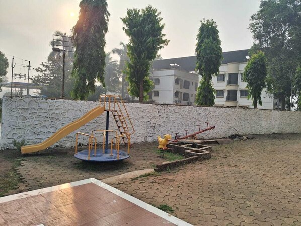 A playground with a slide and climbing structure, surrounded by a paved area and a stone wall. - HOTEL SARIKA (Murbad)
