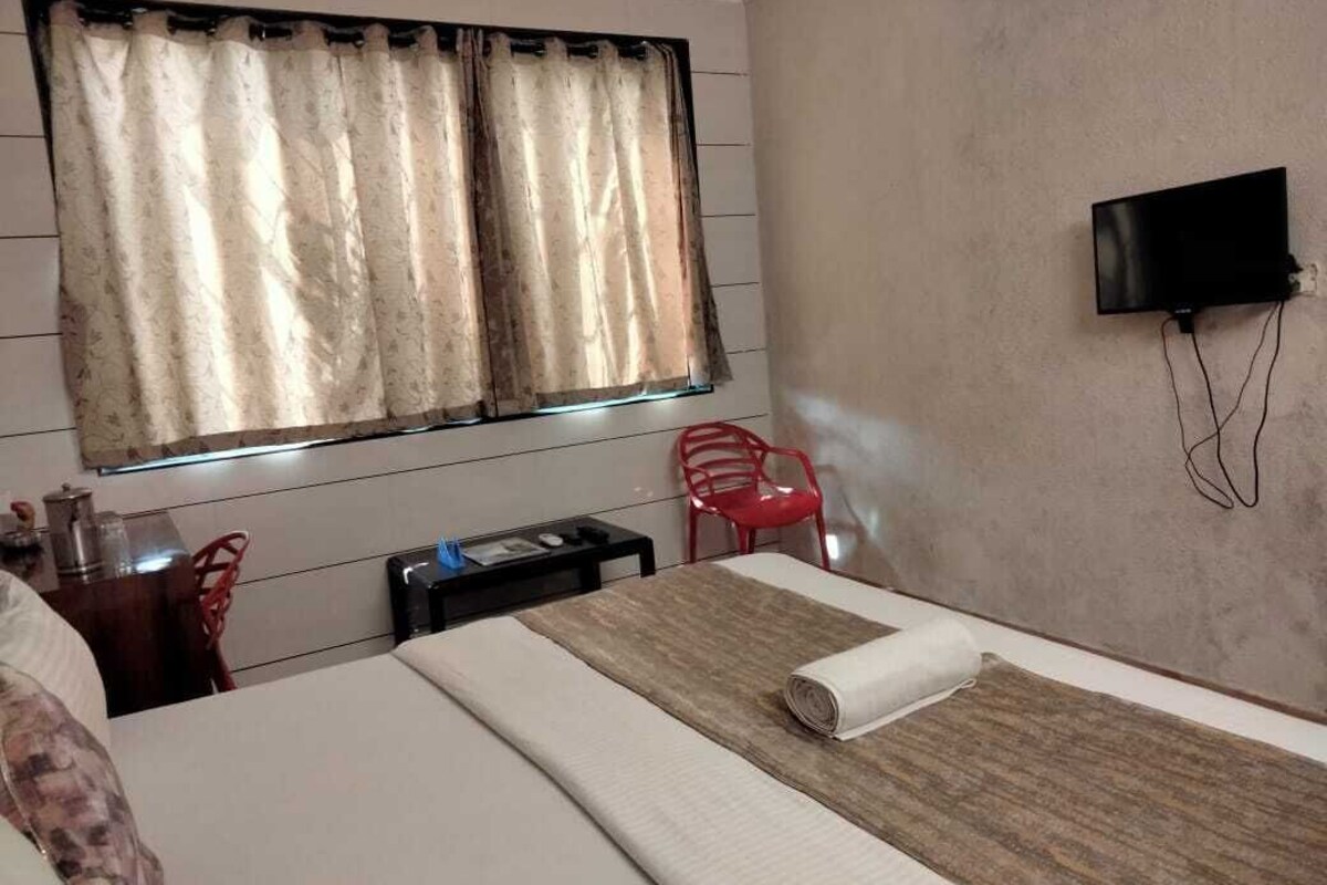 A hotel room with a bed, a TV mounted on the wall, a window with curtains, and a red chair.