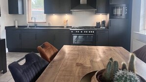 A modern kitchen with dark cabinets, a wooden dining table, and a stove. - Luxurious country house with whirlpool, sauna and private swimming pool (Schoonloo)