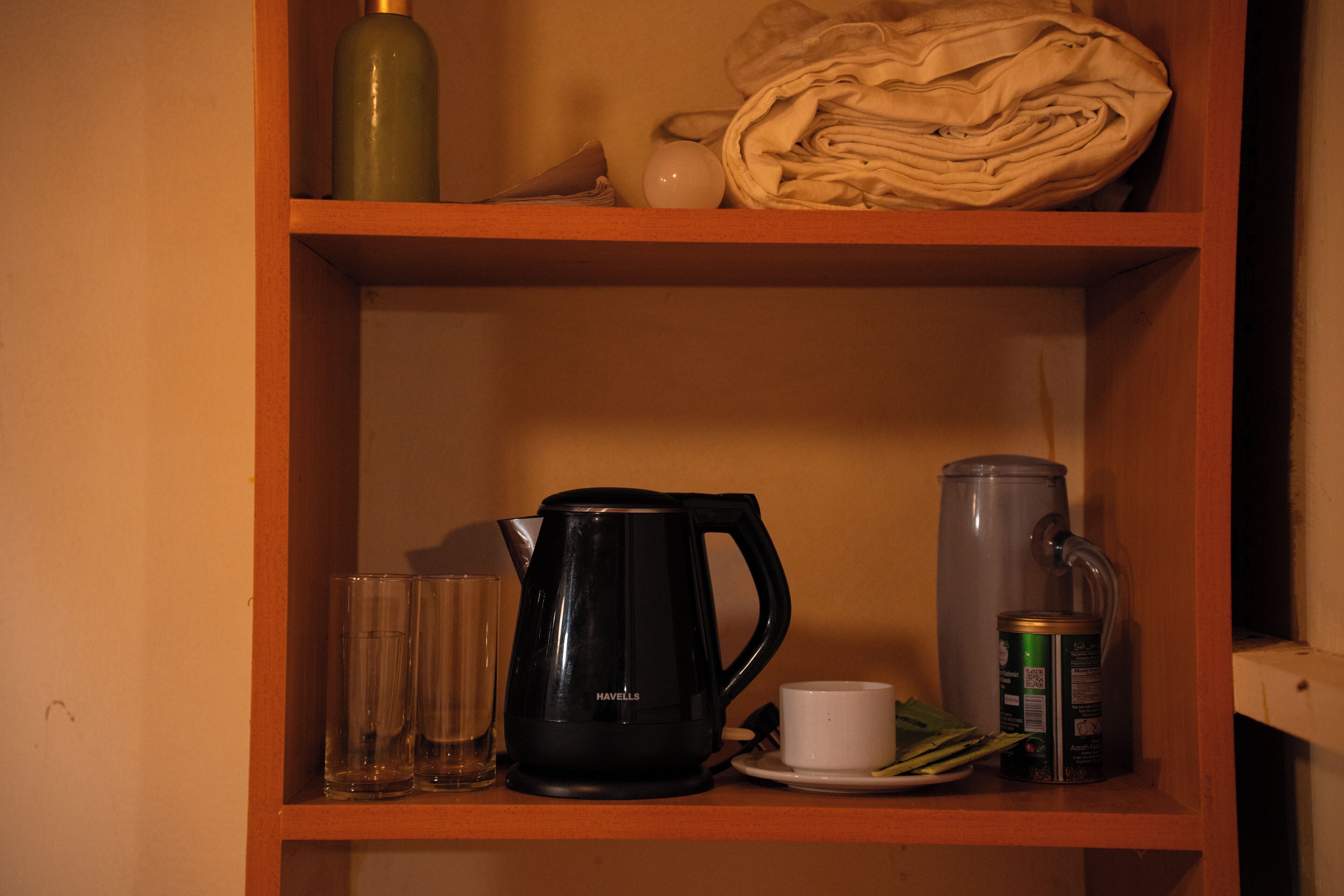 A shelf with a black coffee maker, a glass, a white cup, a rolled-up towel, and a can.