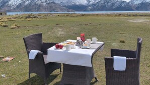 Una mesa de comedor con mantel blanco dispuesta para dos personas, rodeada de sillas de mimbre, con un paisaje montañoso y una masa de agua al fondo.