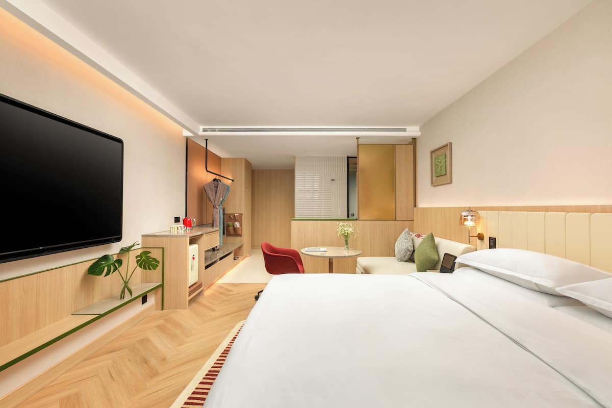 A modern hotel room with a large bed, a flat-screen TV, a small desk with a chair, and a separate seating area with a table and chairs.