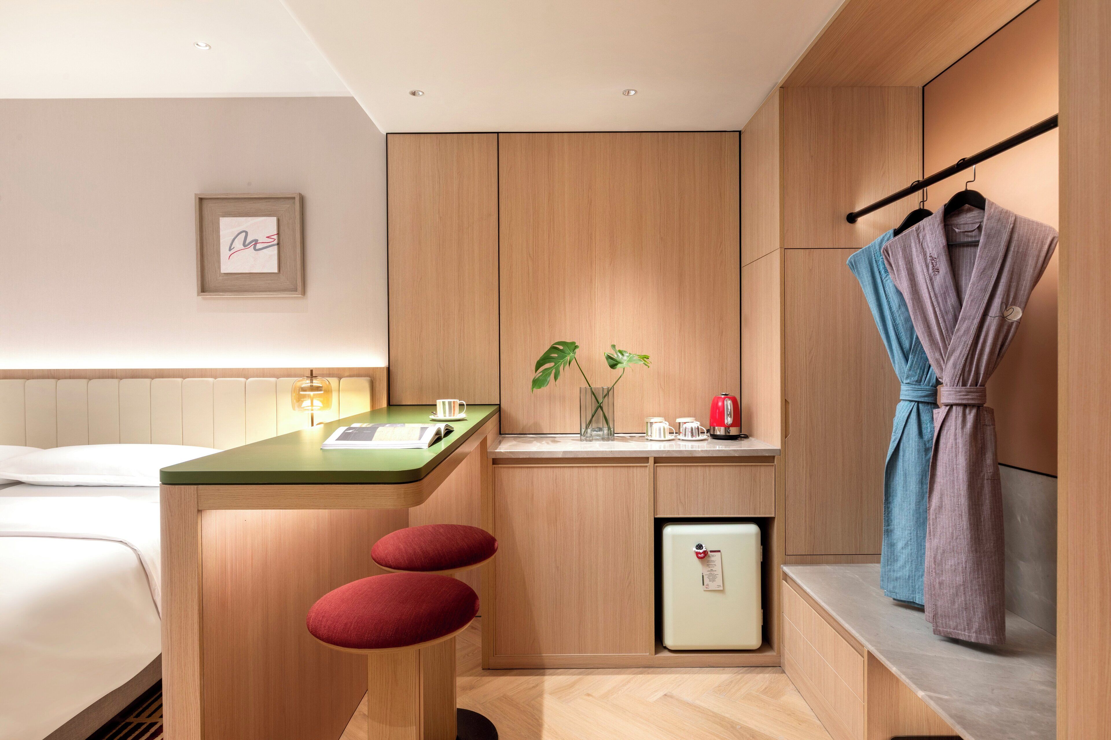 A modern hotel room with a wooden cabinet, a small kitchenette, a bed, and a robe hanging on a rack.