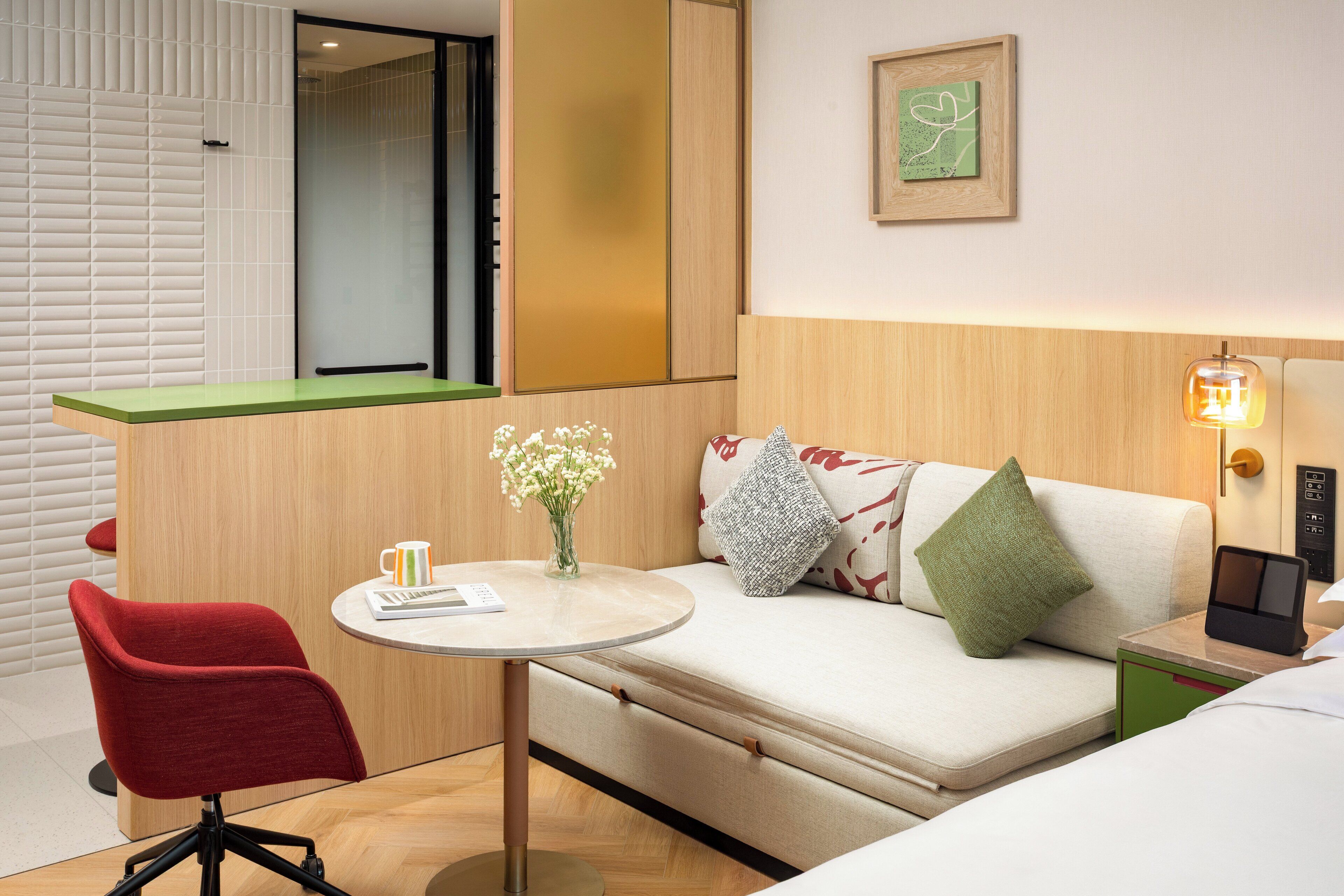 Ein modernes Hotelzimmer mit einer Couch, einem roten Sessel, einem kleinen Tisch mit Blumenvase und einem Nachttisch mit Lampe.